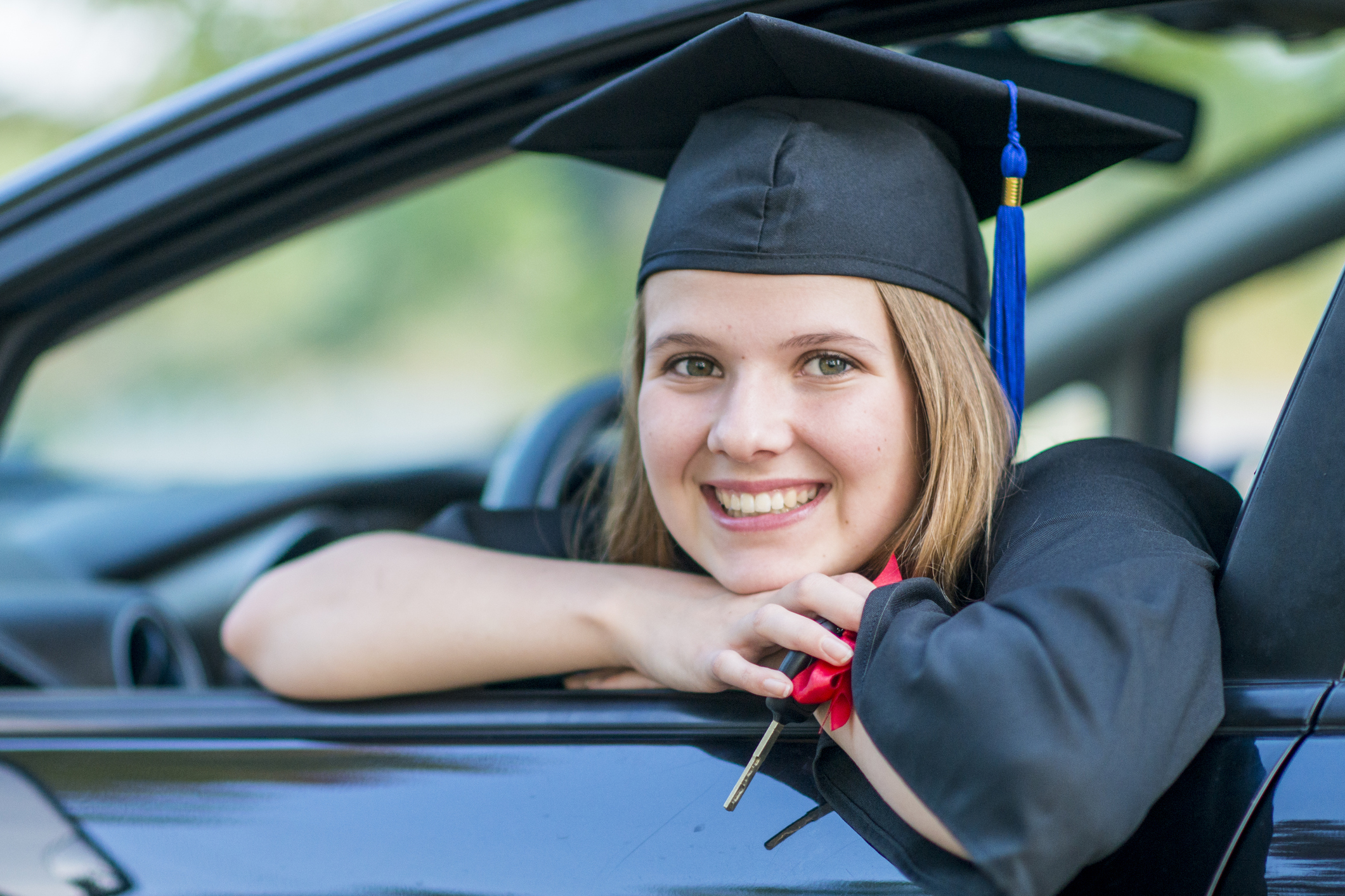 A Caucasian girl is sitting in a new car. She is wearing a graduation robe and cap. She is smiling out the window while holding her keys.